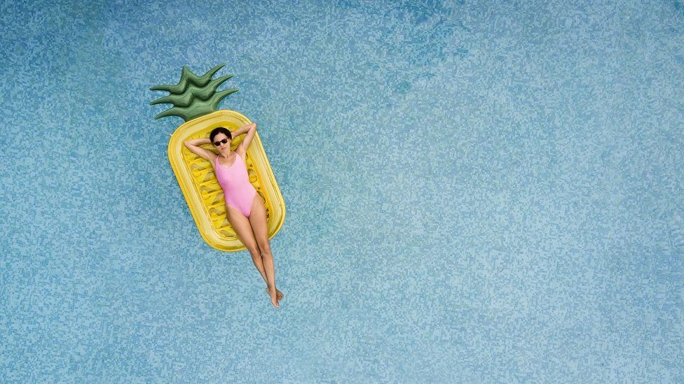 Carefree woman on inflatable pineapple, floating on a beautiful turquoise swimming pool