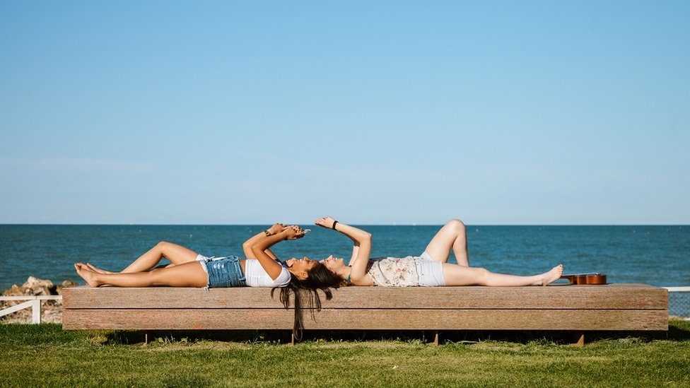 Girls lying down on a bench, by the sea, texting