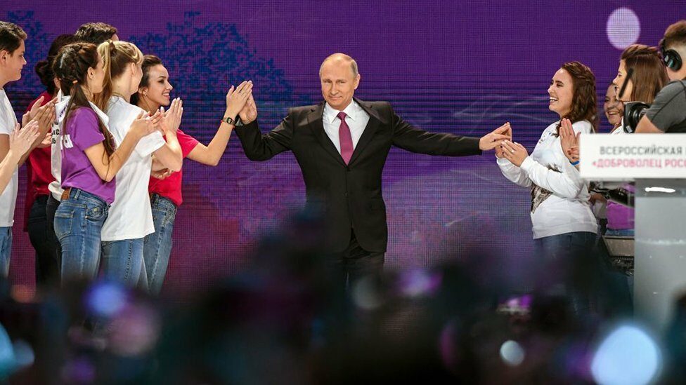 Russian President Vladimir Putin on a stage with women clapping