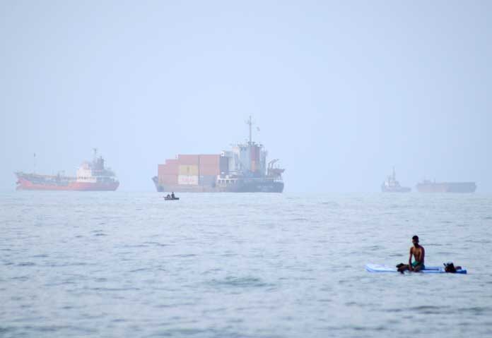 ชายคนหนึ่งนั่งอยู่บนกระดานโต้คลื่น ขณะที่เรือบรรทุกสินค้าและเรือพาณิชย์อื่นๆ จอดทอดสมออยู่ในช่องแคบฮอร์มุซ นอกชายฝั่งเมืองบันดาร์อับบาส ประเทศอิหร่าน เมื่อวันจันทร์ที่ 27 กรกฎาคม 2569