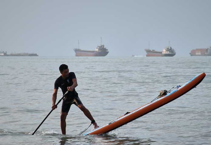 ชายคนหนึ่งกำลังพายกระดานยืนพาย ขณะที่เรือบรรทุกสินค้าและเรือพาณิชย์อื่นๆ จอดทอดสมออยู่ในช่องแคบฮอร์มุซ นอกชายฝั่งเมืองบันดาร์อับบาส ประเทศอิหร่าน เมื่อวันจันทร์ที่ 27 กรกฎาคม 2569