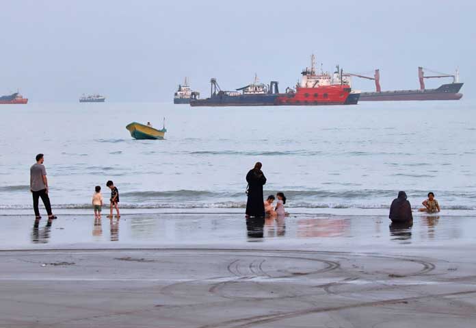 ประชาชนใช้เวลาพักผ่อนที่ชายหาด ขณะที่เรือบรรทุกสินค้าจอดทอดสมออยู่ในช่องแคบฮอร์มุซ นอกชายฝั่งเมืองบันดาร์อับบาส ประเทศอิหร่าน เมื่อวันพุธที่ 5 สิงหาคม 2569